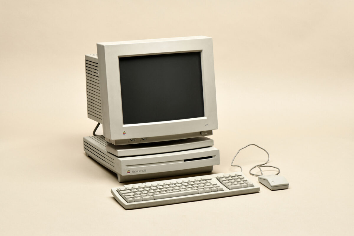 A vintage Apple computer with a CRT monitor, keyboard, and a single-button mouse is displayed against a plain background. The equipment has a beige color typical of 1980s-1990s computer hardware.