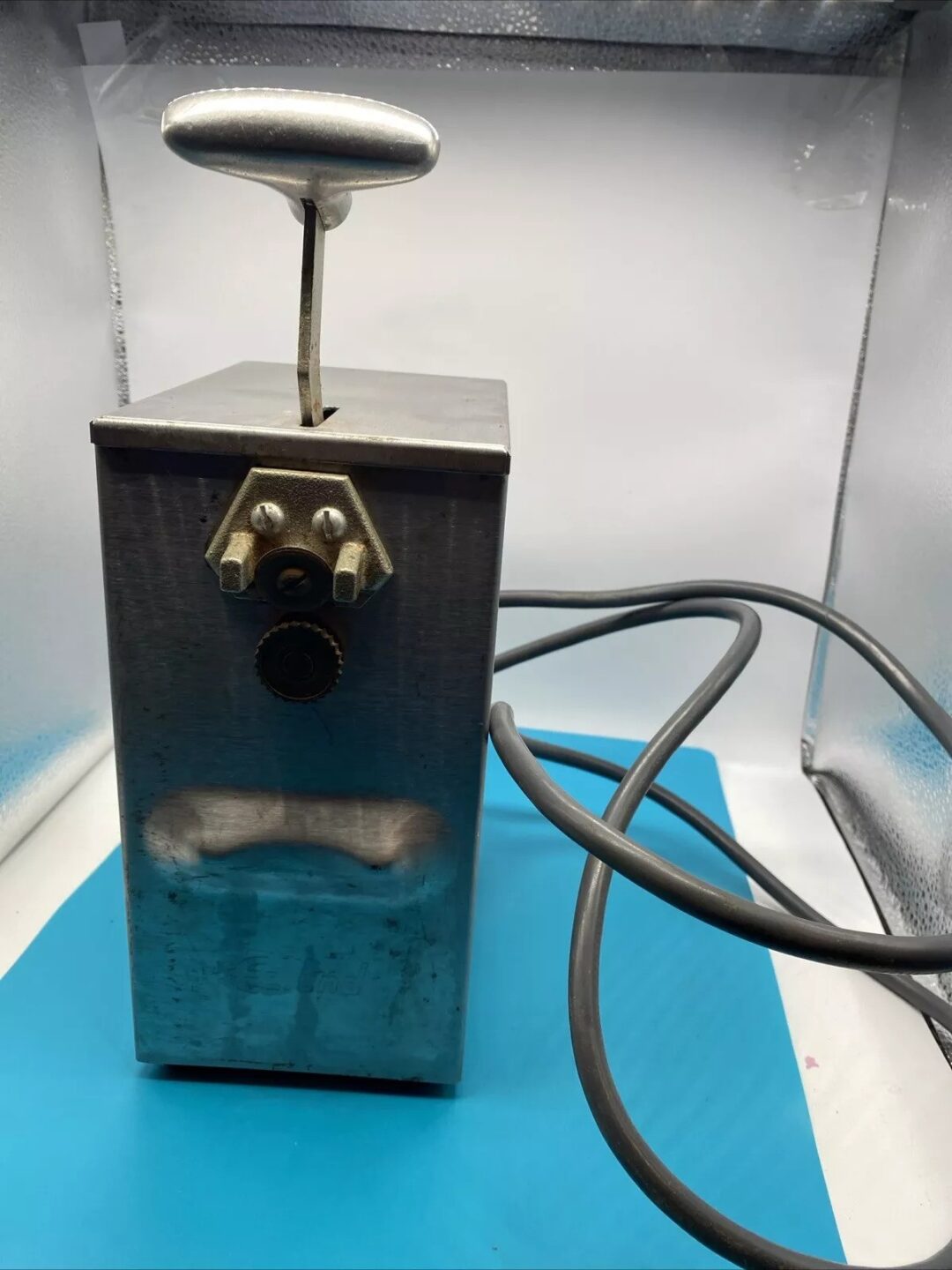 A vintage metal box appliance with a lever handle on top and a power cord extending from its base. It is placed on a blue surface and set against a white and silver background.