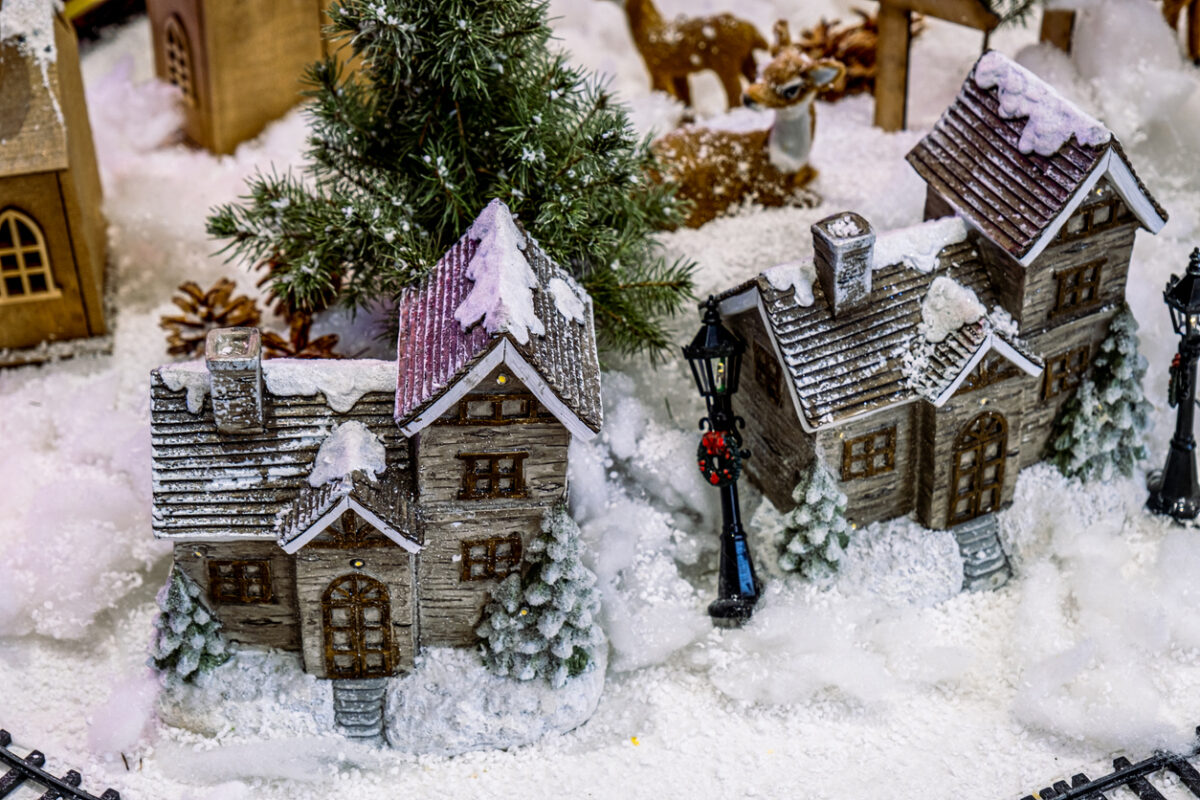 A festive Christmas village scene features two snowy, wooden houses surrounded by pine trees and lampposts adorned with wreaths. Snow blankets the ground, creating a cozy, wintery atmosphere.