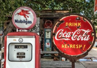 Vintage Mobilgas pump with a red Pegasus logo stands beside a weathered, round Coca-Cola sign that reads "Drink Coca-Cola Refresh." The scene has a nostalgic, mid-20th-century feel, set under a leafy tree.
