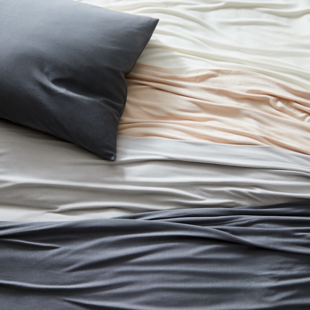 A soft fabric surface with folded and draped layers in shades of gray, beige, and white. A dark gray pillow rests on the upper portion, casting subtle shadows over the gentle undulations of the fabric.