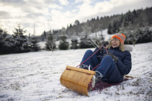 A woman is joyfully sledding on a snowy hillside, wearing a winter coat, gloves, and an orange hat with pom-poms. The background features snow-covered trees and a cloudy sky.