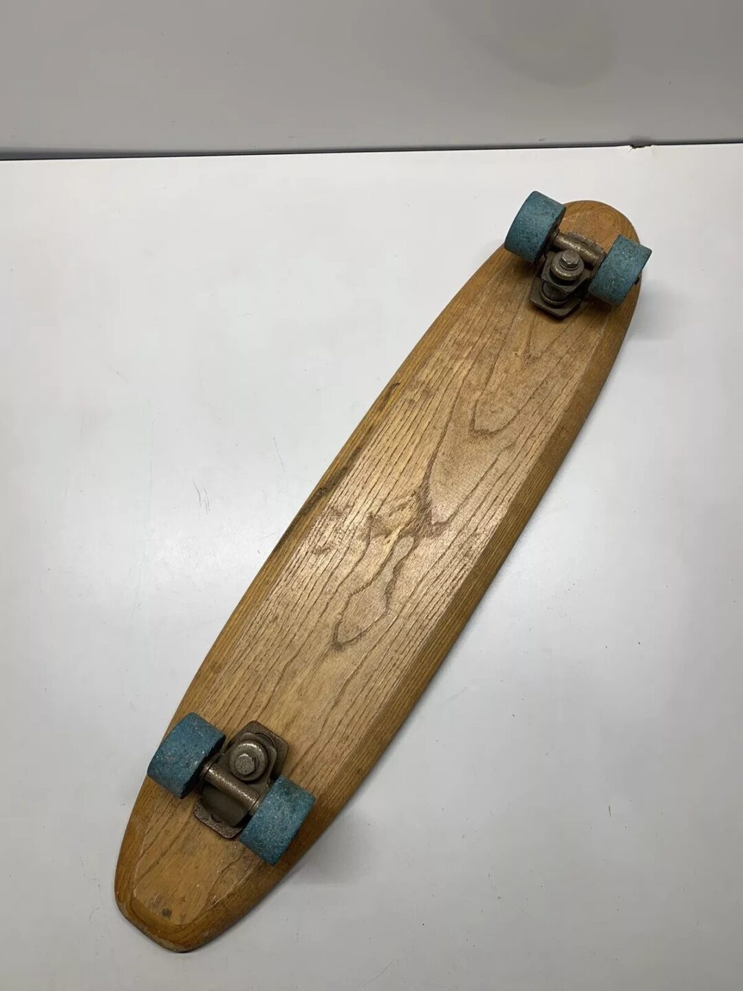 A vintage wooden skateboard with a simple, elongated deck and blue wheels is placed on a white surface. The skateboard shows signs of wear, giving it a nostalgic and classic look.