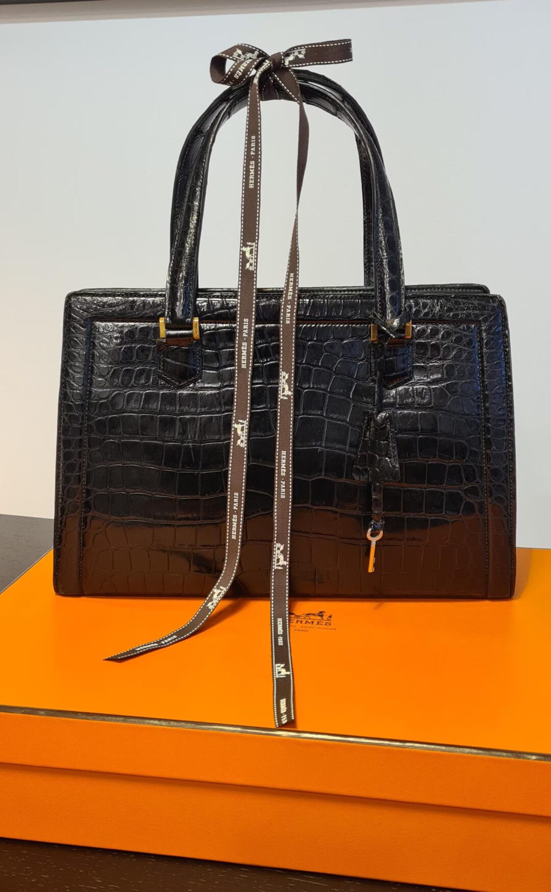 A black crocodile-patterned leather handbag with a structured design sits on an orange box. The bag features gold hardware and a decorative brown ribbon tied in a bow around the handles.