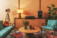 A stylish vintage living room with teal chairs, a circular wooden coffee table with flowers, a wooden sideboard with a record player and speakers, a lamp, a retro camera on a tripod, and a leafy plant.