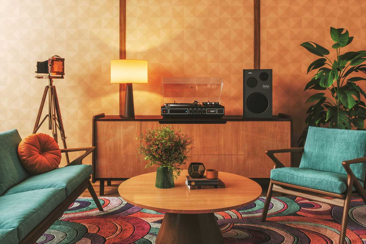 A stylish vintage living room with teal chairs, a circular wooden coffee table with flowers, a wooden sideboard with a record player and speakers, a lamp, a retro camera on a tripod, and a leafy plant.