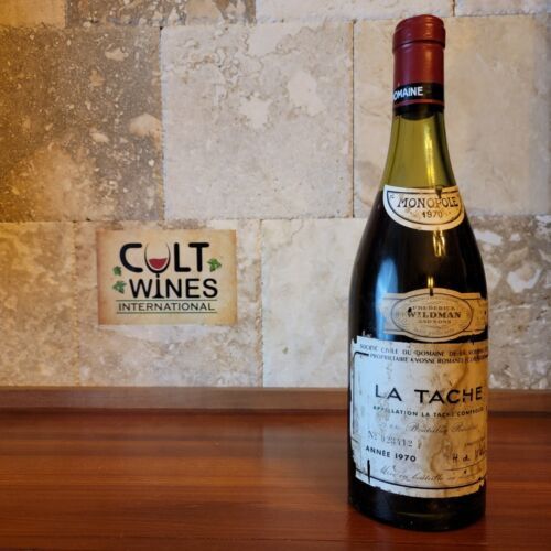 A bottle of 1970 La Tâche wine is displayed on a wooden surface against a textured stone wall. Next to the bottle is a sign that reads "Cult Wines International." The wine label shows its vintage and origin details.