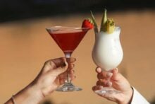 Two hands holding cocktails: a red drink in a martini glass garnished with a strawberry, and a creamy white drink in a curved glass garnished with pineapple and leaves, against a blurred outdoor background.
