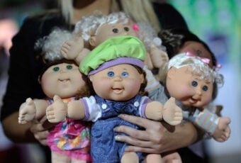 A person holding several Cabbage Patch Kids dolls. The dolls have soft bodies and round faces, each with different outfits and hair styles. The central doll wears a green cap and denim overalls.