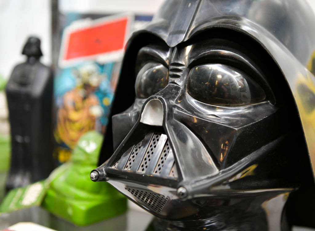 Close-up of a Darth Vader helmet with a reflective surface. In the background, there are blurred vintage comic books and related memorabilia, including another Vader figurine and green objects.