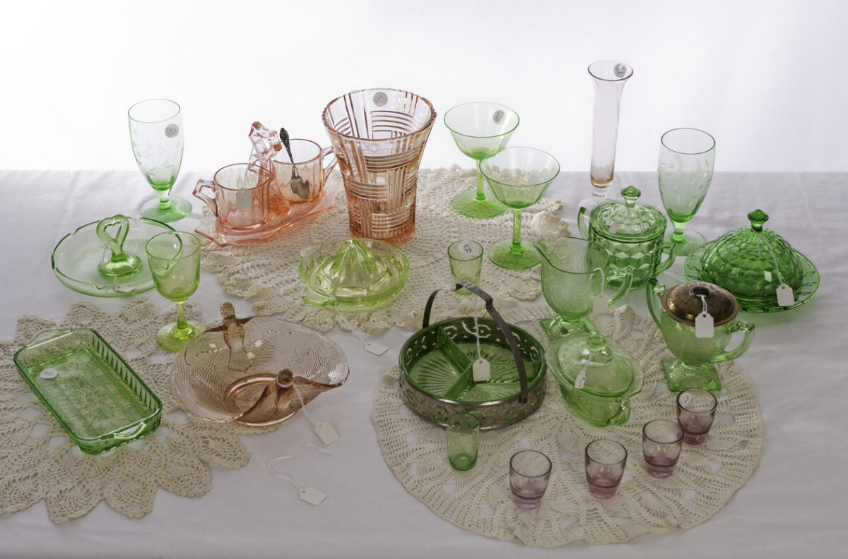 A collection of vintage glassware in green and pink hues displayed on a white lace tablecloth. Various items include pitchers, bowls, plates, and glasses, each with a small tag. The scene is softly lit from above.