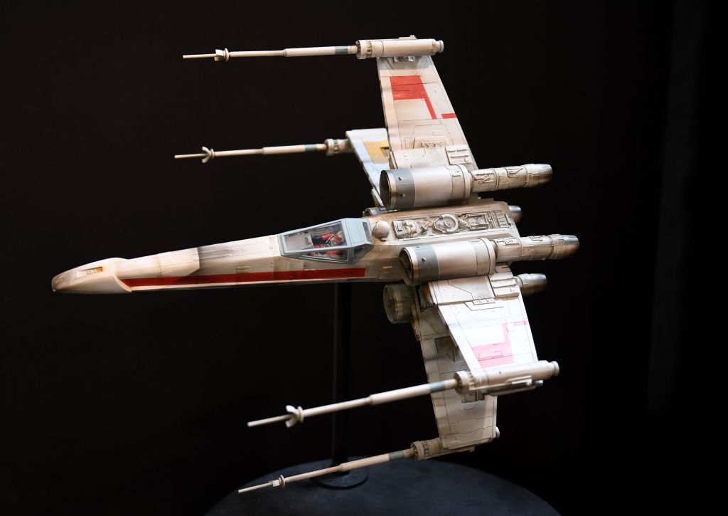 A detailed model of a fictional futuristic spacecraft with a white body and red markings, displayed on a stand against a dark background. The vessel features distinctive wing structures and cylindrical engines.