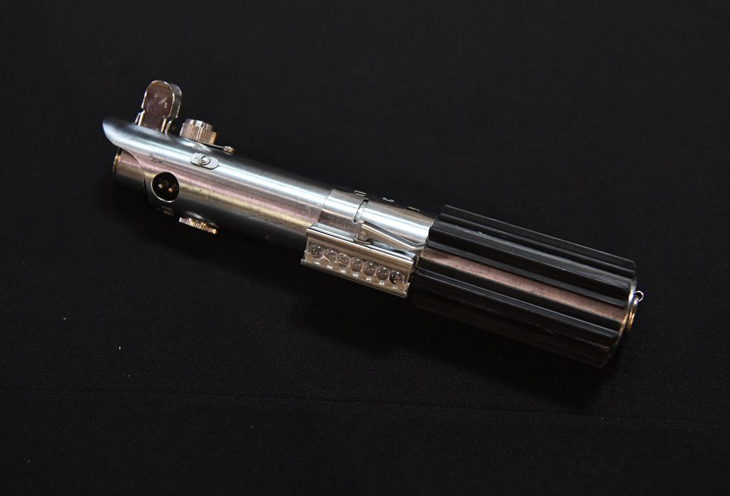 A metallic and black cylindrical object resembling a lightsaber hilt is placed on a dark surface. It has various buttons and details along its length, giving it a futuristic appearance.