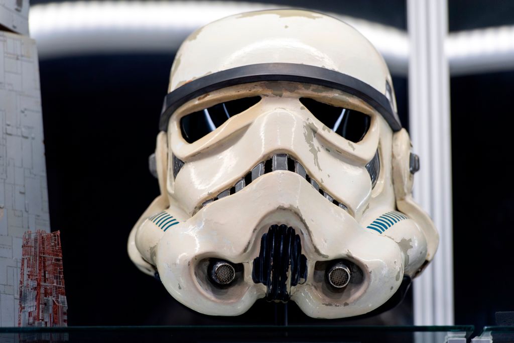 A white and slightly worn stormtrooper helmet with black accents is displayed on a shelf. The helmet features iconic vents and eye slits, with a background of an illuminated surface and a structure to the left.