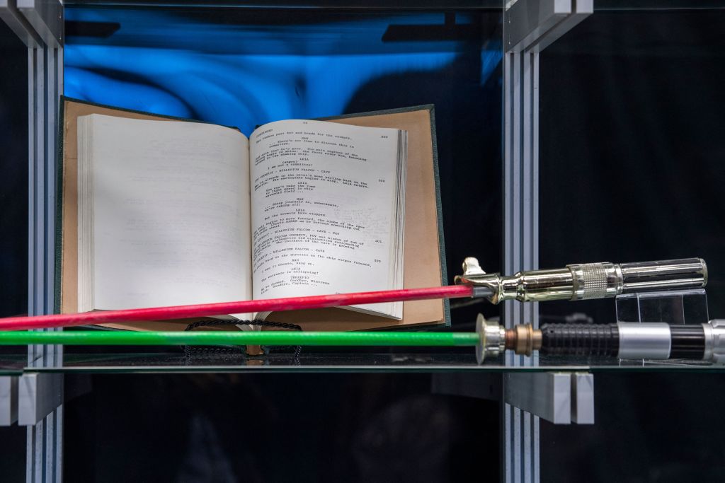 An open book with a script displayed on a shelf, with red and green prop lightsabers positioned in the foreground. The scene is set in a glass display case, reflecting a mix of modern and classic movie memorabilia.