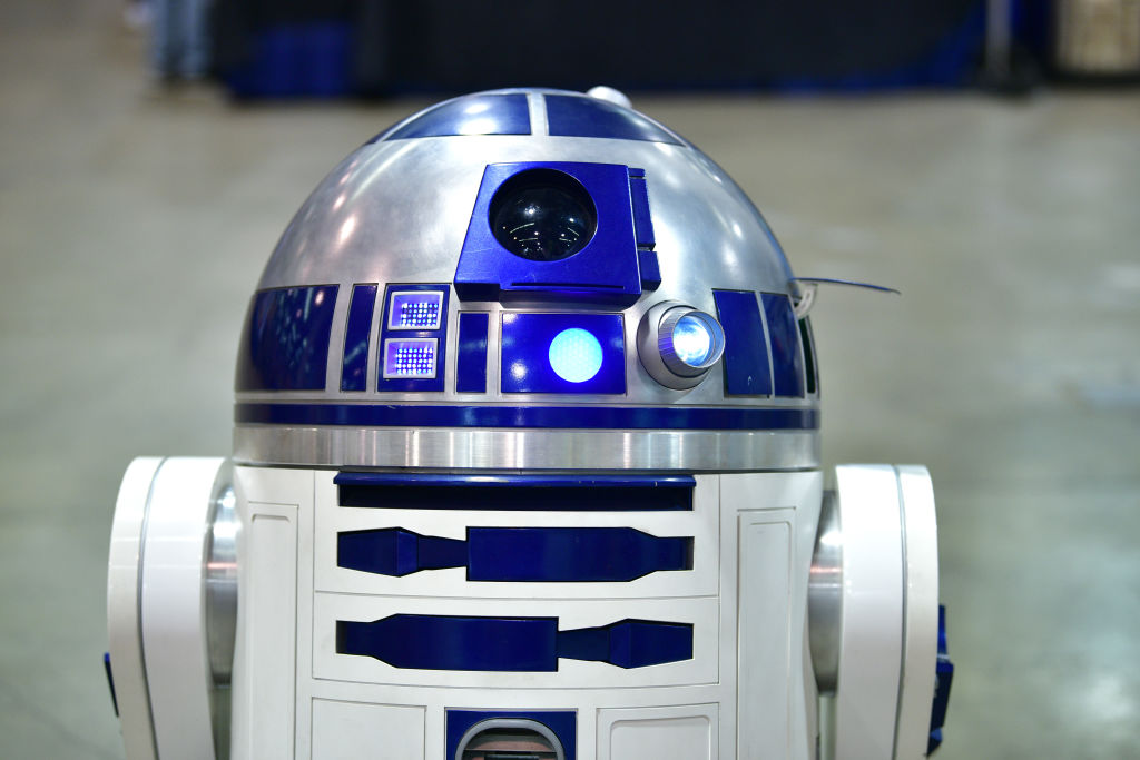 A close-up of a blue and silver robot with a dome-shaped head and cylindrical body, resembling a character from science fiction. The robot has various lights and panels on its surface, set against a blurred, neutral background.