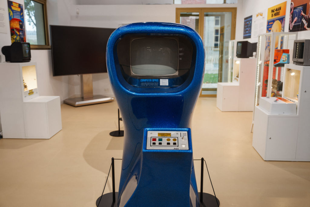 A vintage arcade game machine with a blue exterior is displayed in a museum setting. The machine has a small screen and a control panel with buttons and a joystick. The room features other retro technology exhibits and a large screen in the background.