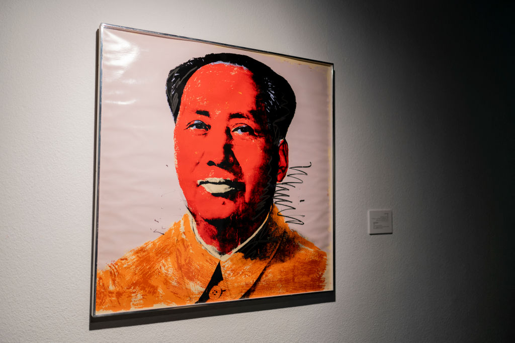 A framed painting depicting a stylized portrait of a man with an orange jacket and red face set against a light background, hanging on an art gallery wall with lighting from the left.