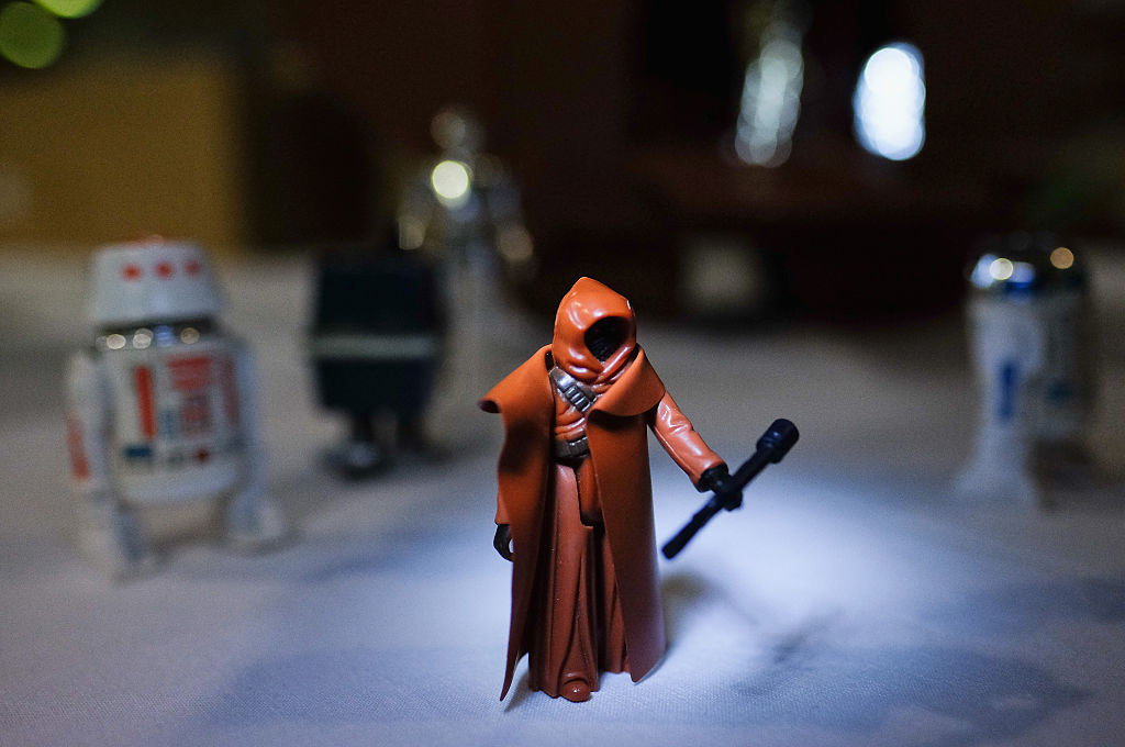 A hooded orange figure, holding a staff, is illuminated under a spotlight on a table. Surrounding it are blurred figures of small robot toys in the background. The setting is dimly lit, enhancing the mysterious ambiance.