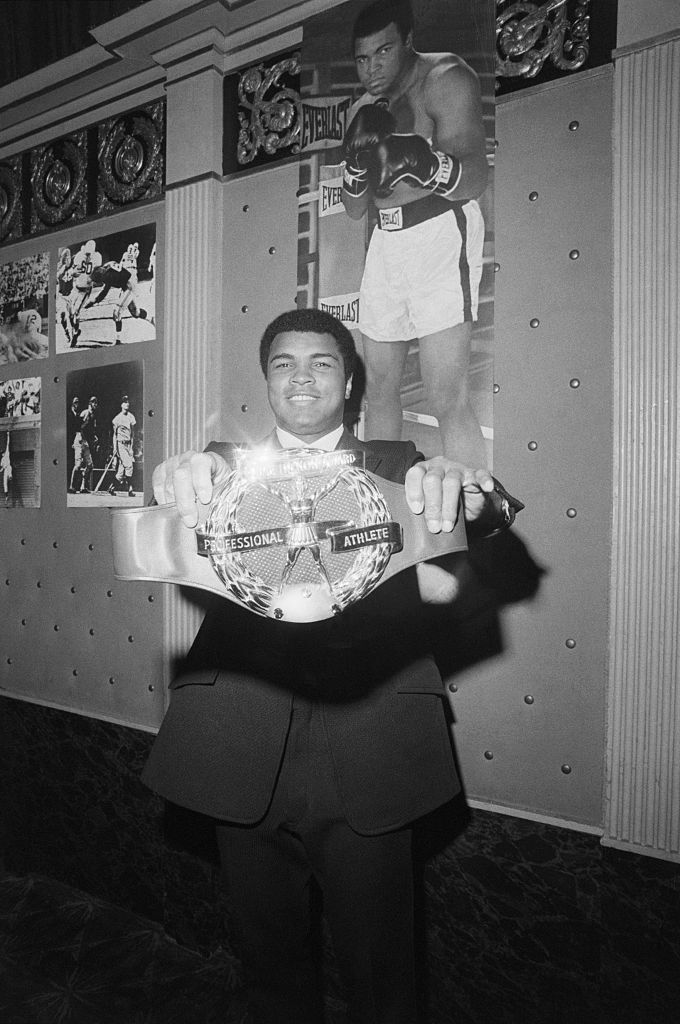 A man in a suit holds a championship belt with "Professional Athlete" inscribed. Behind him, a large poster and photos of a boxer in action are displayed on the wall.