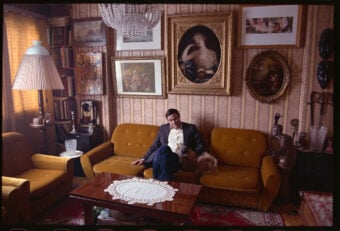 A person sits on a vintage mustard-yellow sofa, petting a dog in a living room filled with eclectic art and ornate decor. The walls display various framed paintings, and the room features a large chandelier and a decorative lace runner on the table.