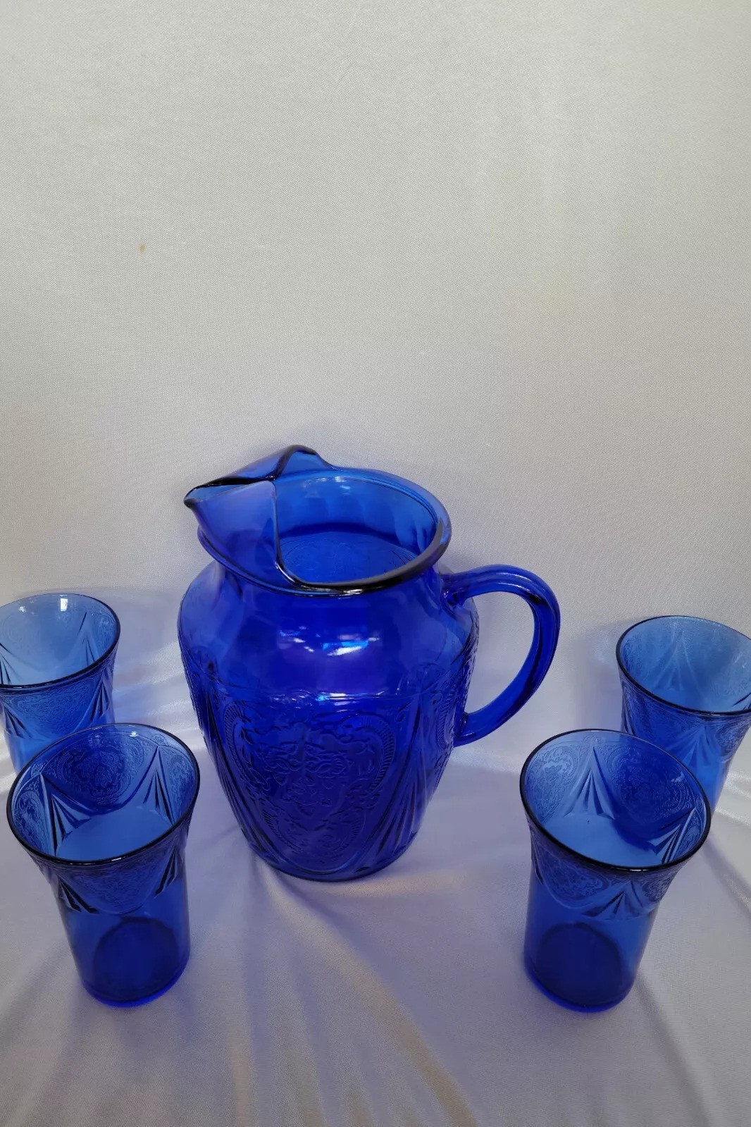 A blue glass pitcher with a decorative design is surrounded by four matching blue glass tumblers, all set against a plain white background.