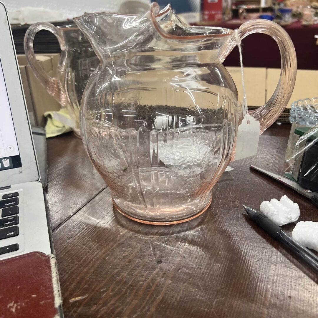 A pink glass pitcher with a vintage design sits on a wooden table. It has a curved handle and a wide spout. Nearby are a laptop, a pen, and decorative items.