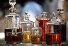 A variety of elegant, transparent glass bottles with ornate stoppers contain different shades of liquid, ranging from amber to golden yellow. The bottles are arranged on a reflective surface with a blurred background.