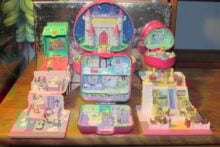 A collection of Polly Pocket playsets displayed on a wooden surface. The sets include a castle-themed compact and various dollhouse-style compacts with colorful, detailed interiors, featuring small figures and accessories.