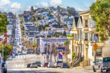 A vibrant city street scene with a steep hill. Cars line the road, flanked by colorful, varied architecture. Trees dot the sidewalks, and multi-story buildings and houses rise in the distance under a blue sky with fluffy clouds.