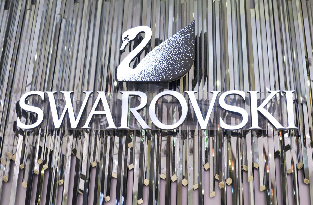 Swarovski logo displayed on a reflective wall, featuring a stylized swan above the brand name in elegant, bold letters. The background has a metallic, shimmering texture.