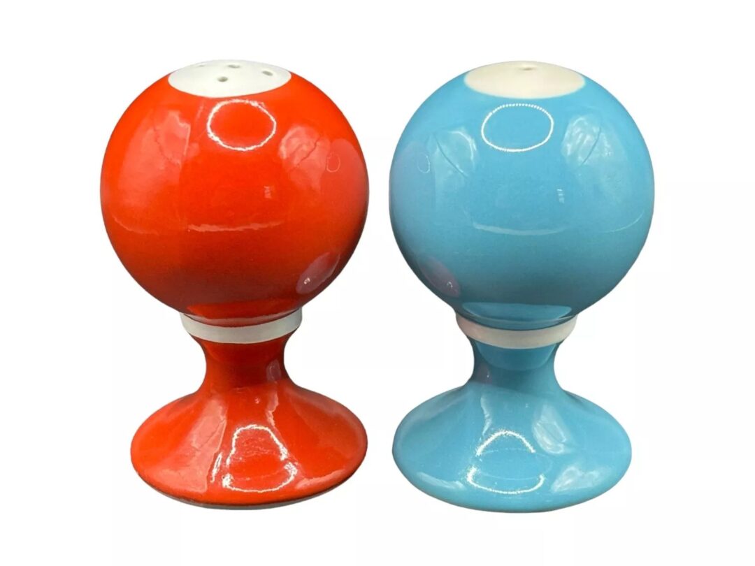 Two round, glossy salt and pepper shakers on pedestal bases. The left one is red, and the right one is blue, each with a white top with small holes. The background is plain white.