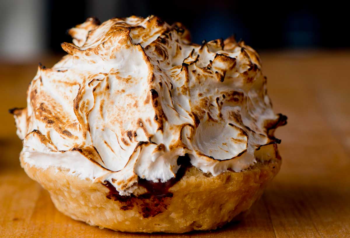 A close-up of a baked Alaska dessert featuring toasted, golden-brown meringue on top of a flaky pastry crust, set on a wooden surface.