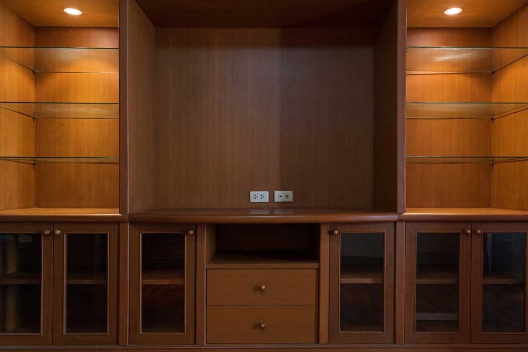 Built-in wooden entertainment center with glass shelves and cabinets, two drawers in the center, and a large empty space for a TV. Two electrical outlets are visible on the back panel, with overhead lights illuminating the shelves.