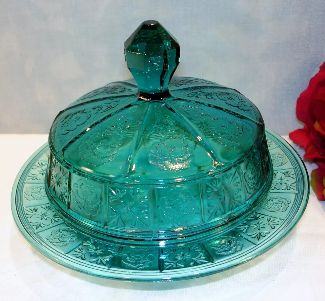 A vintage green glass butter dish with an ornate patterned lid and a matching base. The lid features a faceted knob handle. A red flower is partially visible on the right side.