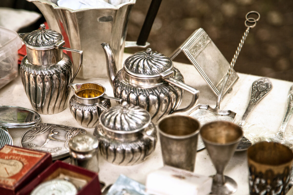 An assortment of vintage silverware, including teapots, cups, and trays, displayed on a table. The pieces have an ornate, ribbed design. The table has additional small items, hinting at a market or antique fair setting.