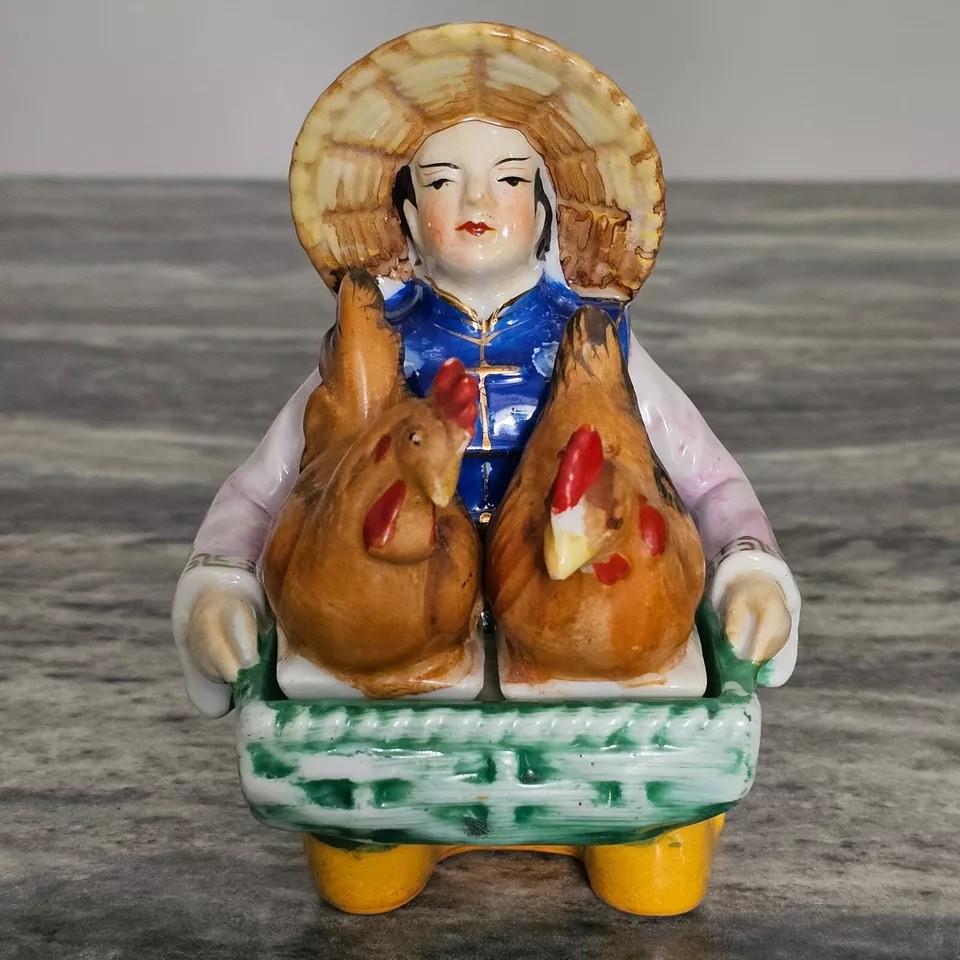 A ceramic figurine of a person wearing a straw hat and blue vest, holding two brown chickens. The figure is seated with a green base, set against a gray marbled background.