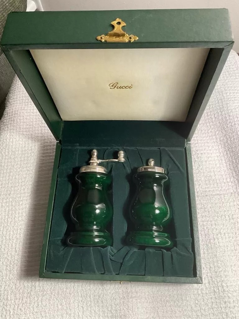 A green Gucci box containing a set of two elegant dark green salt and pepper shakers with silver tops, nestled in a custom velvet-lined compartment. The box lid displays the Gucci logo.