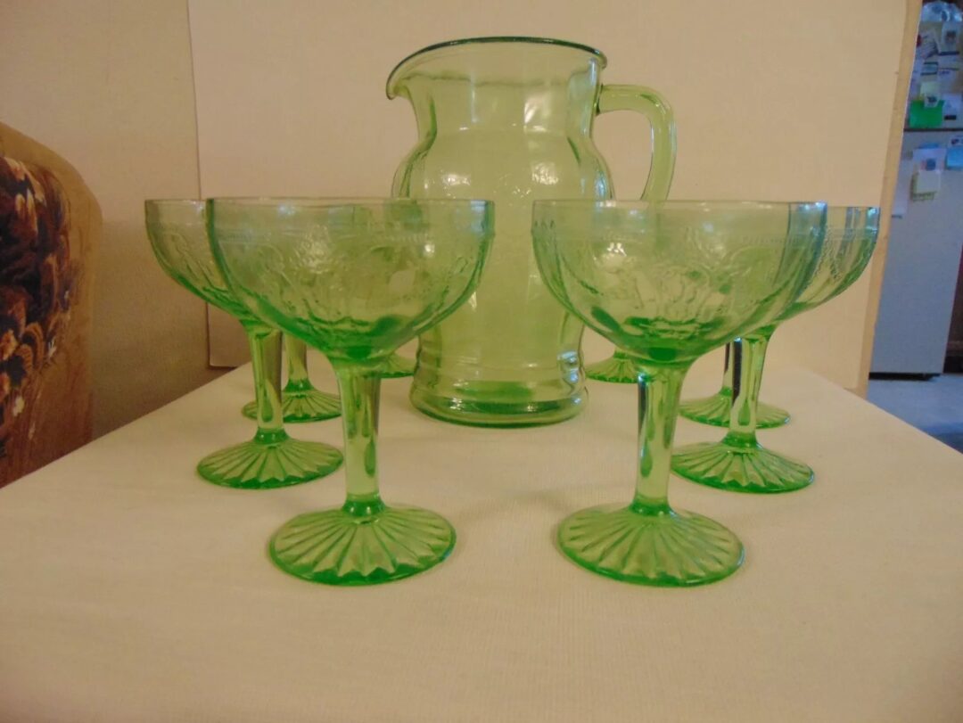 A set of vintage green glassware on a tabletop, featuring a large pitcher and six matching stemmed glasses arranged in a semicircle in front of it. The glassware has an ornate, embossed design.