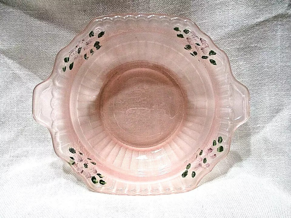 A pink, transparent glass bowl with scalloped edges and handles. It features a floral design with green leaves and small pink flowers around the rim, set against a light fabric background.