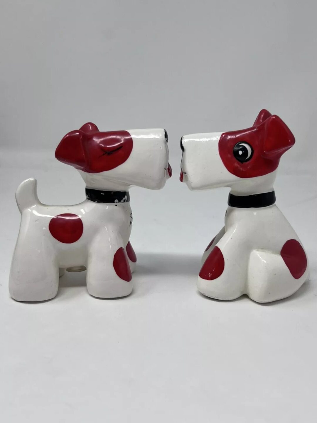 Two ceramic dog figurines with red spots facing each other on a white background. Each has a black collar and similar facial features, appearing to touch noses.