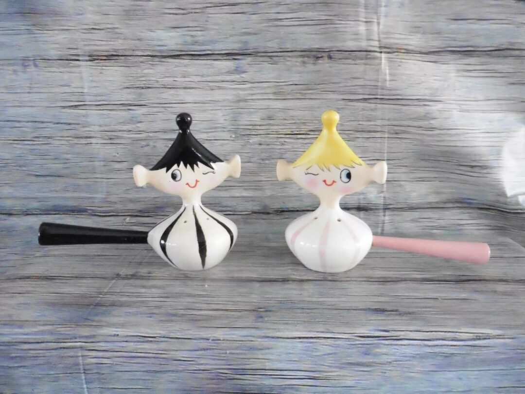 Two cute, smiley character salt and pepper shakers with painted black and yellow hair stand against a rustic wood background. The shaker on the left is wearing a black tie, while the one on the right has a pink tie.