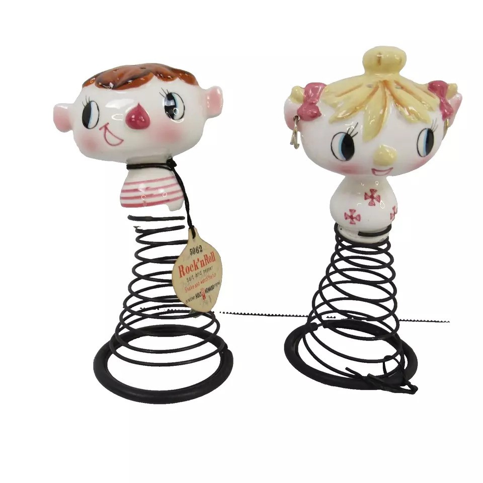 Two whimsical bobblehead figures on spring bases. One has red hair, rosy cheeks, and a striped shirt. The other has blonde hair with pink bows and a floral dress. Both have large eyes and are designed in a playful, artistic style.