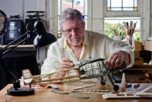 A man in a light shirt is building a model airplane at a wooden table. He holds a small tool to work on the plane's fuselage. The room has stained glass windows, and various tools and parts are scattered across the table.