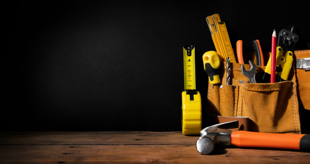 A leather tool belt with various tools, including a tape measure, screwdrivers, pliers, and a hammer, is placed on a wooden surface against a dark background.