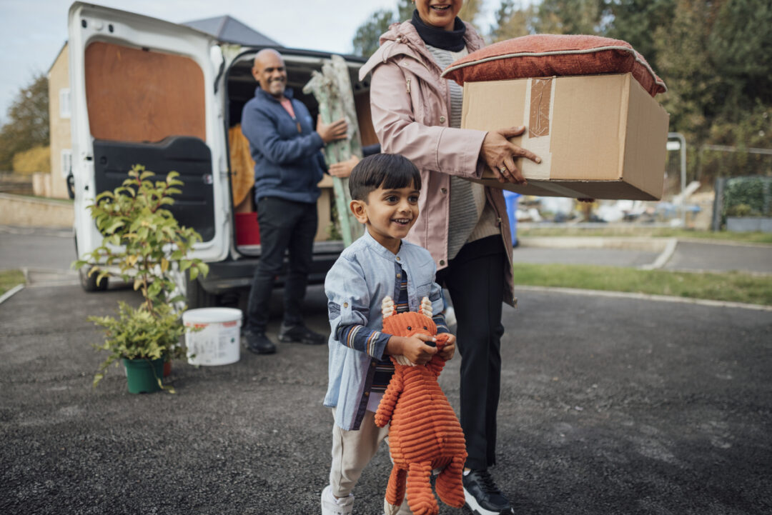 A child holding a plush toy walks happily in front of an adult carrying a cardboard box and a cushion. In the background, another adult unloads items from a van on a driveway. Outdoor scene with greenery.