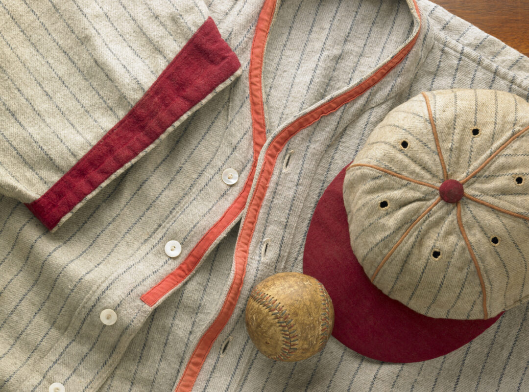 A vintage baseball jersey and cap are laid out on a wooden surface. The jersey is light with dark stripes and features red accents, including a patch and collar. A worn baseball rests on the jersey. The cap has a similar striped pattern.