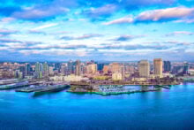 Aerial view of a city skyline with numerous skyscrapers under a partly cloudy sky. In the foreground, a large body of water with a docked military ship and marina can be seen.