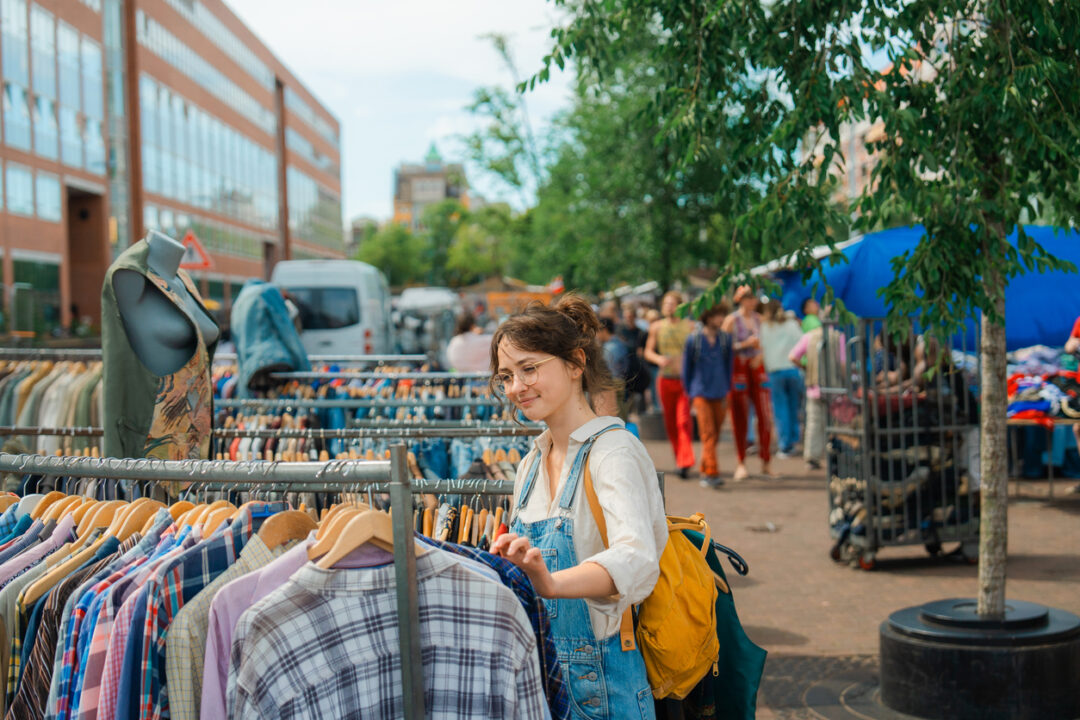 A person with glasses and a yellow backpack browses clothes at an outdoor thrift market. They are surrounded by racks of shirts. Other shoppers and vendors are visible in the background under trees and a blue umbrella.