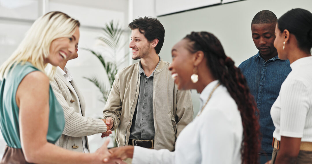 A group of five people stand in an office setting, engaging in conversation and shaking hands. A woman with blonde hair and a woman with braids are in the foreground. A man with a jacket and two others are in the background.